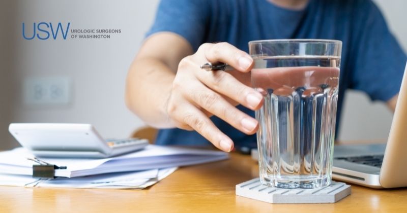Man in blue tee shirt sits at desk reaching for glass of water as he works to stay hydrated for his urological health. Urologic Surgeons of Washington logo at top left.